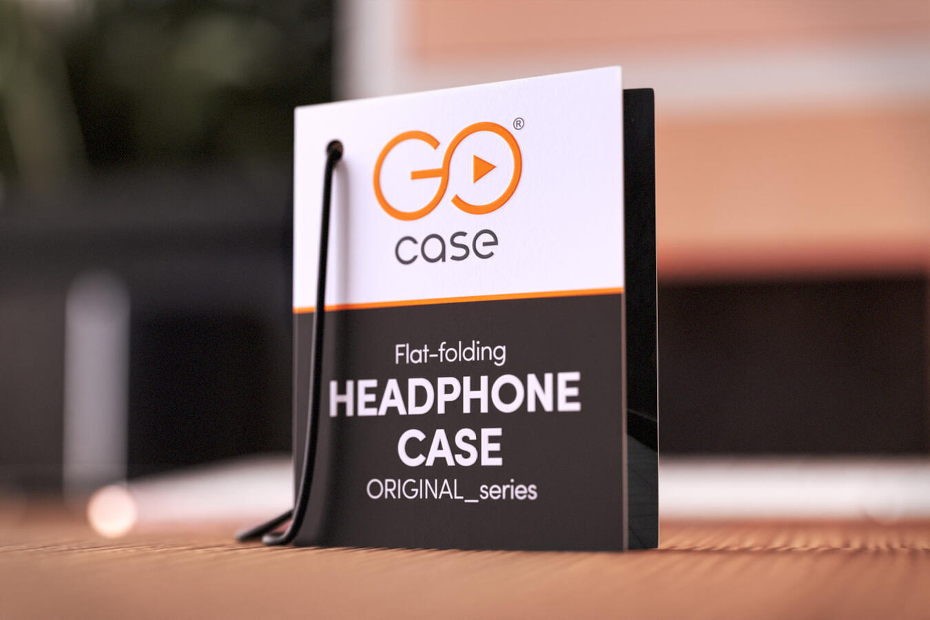 Close-up of GOcase flat-folding headphone case tag, featuring the brand's orange and black logo, resting on a wooden table.