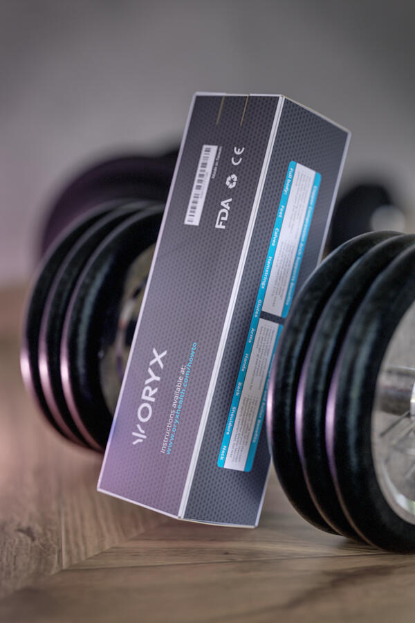 Oryx product box, dark grey with purple accents, detailing FDA, recycling, and CE markings. Propped between two dumbbells on a wooden floor.