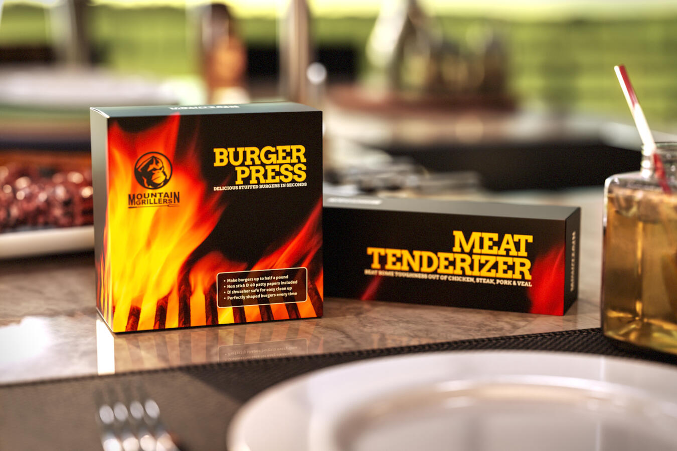 Two Mountain Grillers product boxes, a Burger Press and a Meat Tenderizer, featuring a fiery design, displayed on a countertop.