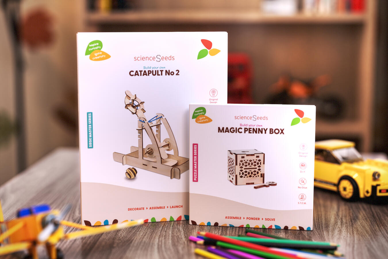 Two scienceSeeds educational toy boxes, a catapult and a magic penny box, displayed on a wooden surface with colorful pencils and toy cars in the background.