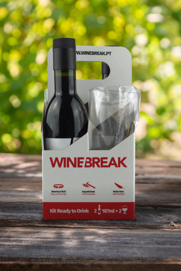 A Wine Break Kit Ready to Drink cardboard carrier containing two small bottles of red wine and two plastic wine glasses, placed on an outdoor wooden table.