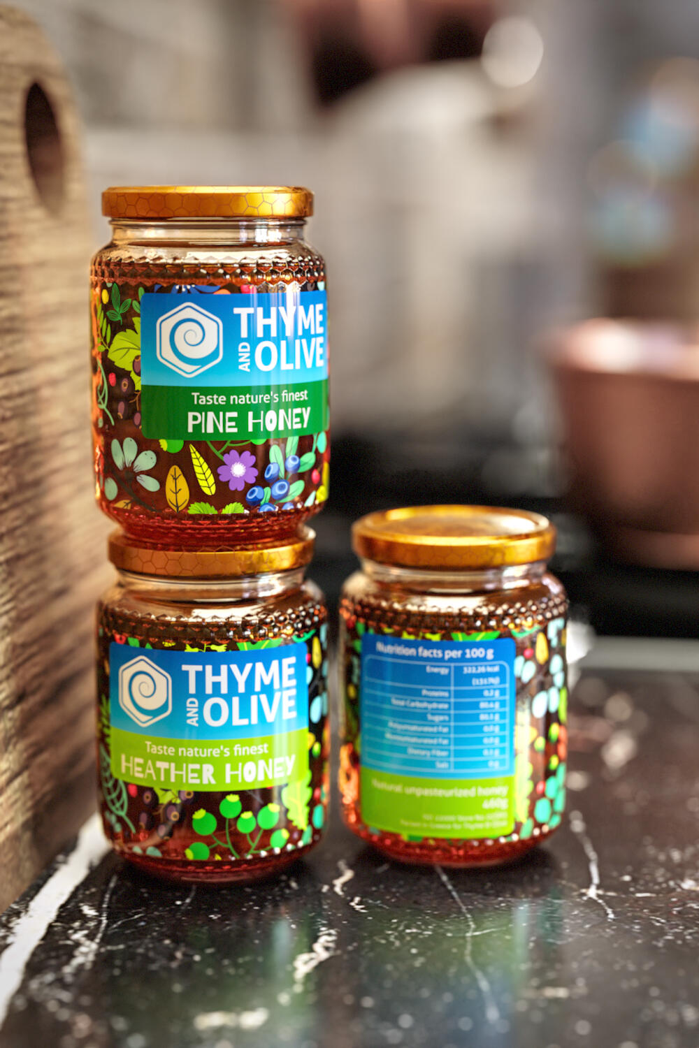 Three Thyme and Olive honey jars with colorful labels, Pine Honey and Heather Honey stacked, and another showing nutritional facts, on a dark kitchen counter.